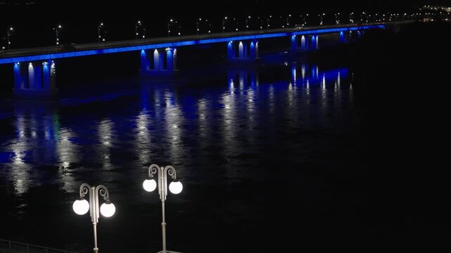 A wide night landscape showing a long bridge spanning a river, vibrantly illuminated with bright blue neon lights reflecting on water.