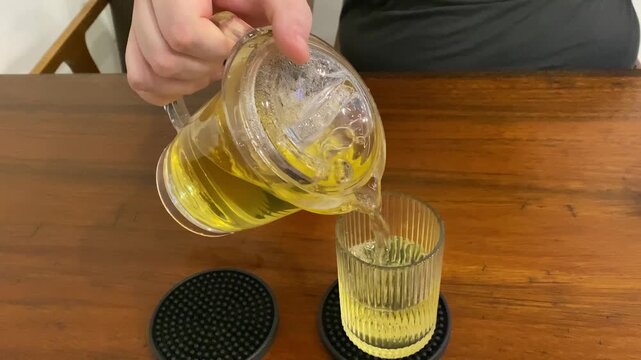 Pouring refreshing green iced tea from glass teapot into ribbed glass cup horizontal video, close up of cold herbal tea beverage on wooden table in cafe