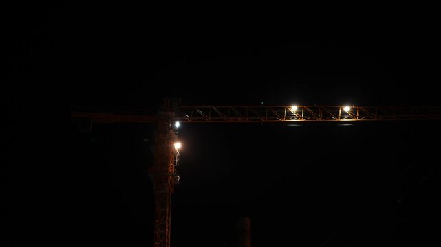 A close-up night view of a tower crane jib illuminated by bright spotlight work lights against a pitch-black sky.