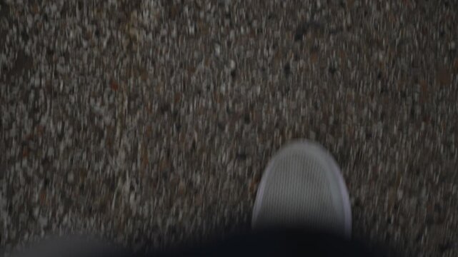 A top-down, motion-blurred view of a person wearing a light grey sneaker while walking on a textured asphalt road surface.