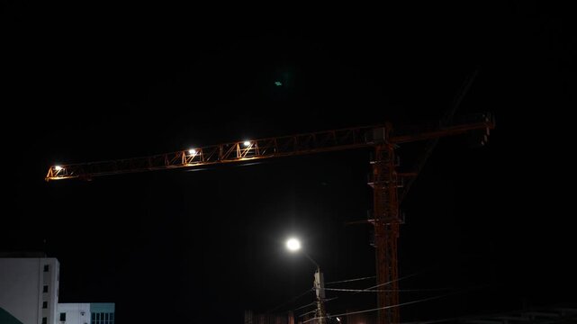 A close-up night view of a tower crane jib illuminated by bright spotlight work lights against a pitch-black sky.