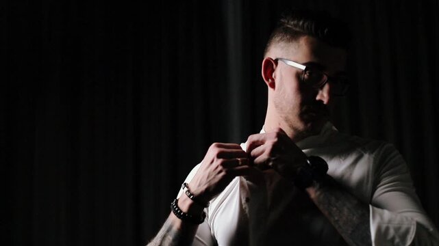 Young man wearing glasses carefully buttons white cotton shirt while standing in front of black draped curtain, both hands engaged in guiding top buttons through placket with fingers pinching fabric