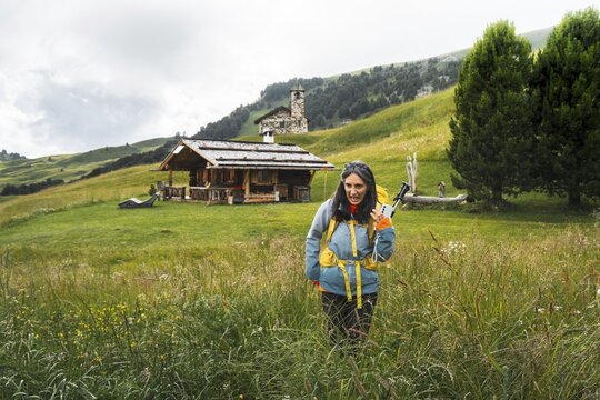 Disgusted woman hiker in waterproof clothing pulling funny face walking facing fears in tall high wet wildflower meadow background Malga Rossalm loop trail outdoor wilderness.Dolomites Italy