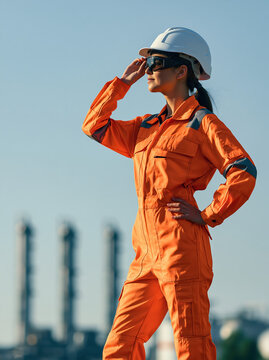 Female industrial safety specialist surveying large facility in orange protective coverall under clear sky