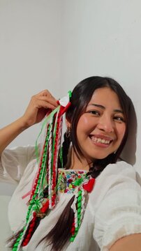 Mexican woman celebrating independence day wearing a traditional embroidered blouse, tricolor hair ribbons, and braids while taking a selfie against a white background