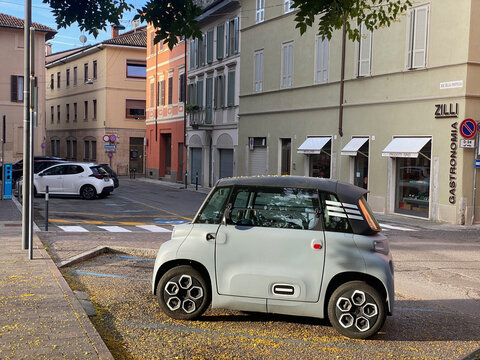 A light gray Citroen Ami electric microcar is parked on a cobblestone street in a European town, surrounded by fallen yellow leaves
