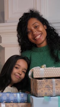 Cheerful african american mother and her cute daughter posing with a pile of wrapped gift boxes, celebrating christmas eve together in a cozy home environment with genuine smiles