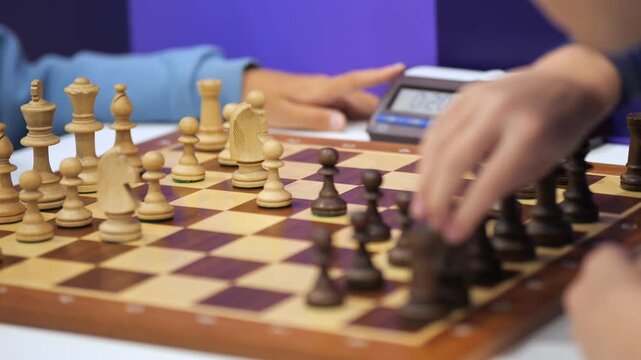 Two people play chess with wooden pieces on a checkered board. One hand moves a black pawn forward. Digital chess clock sits beside the board. The background is purple, blurred, and softly lit