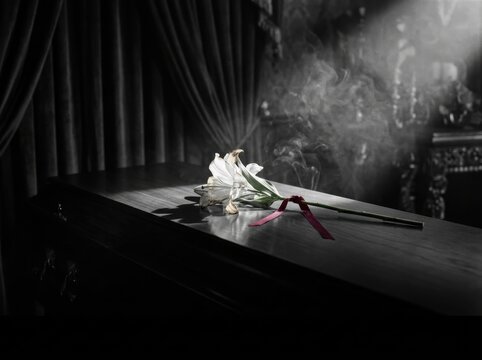 Dramatic noir funeral still life featuring a white lily with crimson ribbon on a polished coffin with cinematic lighting and copy space.