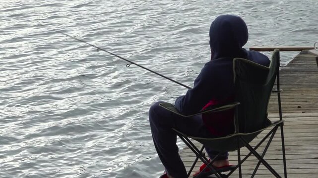 A man sits on a lawn chair on a wooden dock, casting his fishing line out into the calm lake water before slowly reeling the fishing rod back in.