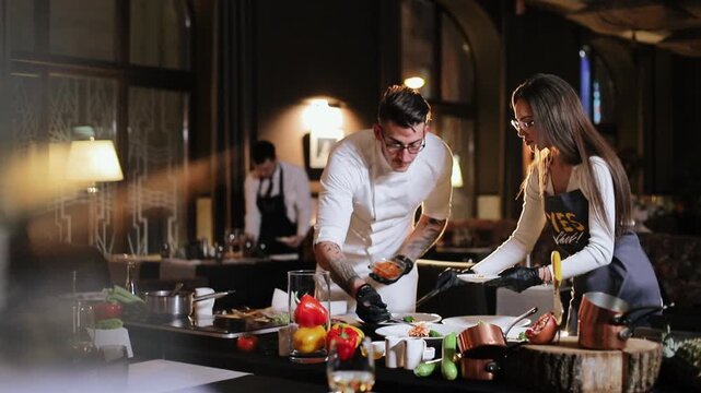 Male chef in white jacket uses gloved hands and utensil to transfer food into bowl while female assistant in apron steadies plate and watches placement, showing coordinated precise plating and