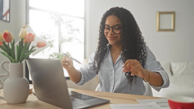 Woman at laptop raises arms and claps smiling in a home office studio with tulips and papers on desk; work success celebration.