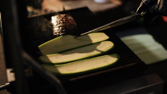 Three zucchini halves arranged lengthwise on heated ribbed grill while metal tongs press upper slices and foil-wrapped round packet at rear releases faint steam, low-angle three-quarter close-up