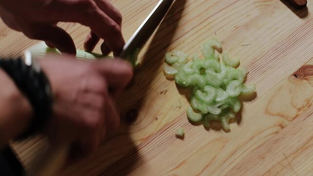 Close-up slightly overhead view of hands preparing pale green celery on light wood cutting board, with celery, knife, hand, cuttingboard detail, showing slice, chopping, chef, stalk context