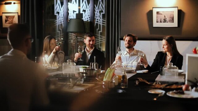 Group of four young professionals seated waist-up at rectangular dining table raise wine glasses in mutual toast, smiling and making eye contact to convey convivial celebration, with dinner