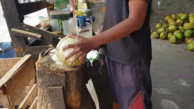 Close up of hands cutting and peeling fresh young coconut using a large knife. Authentic street food preparation, organic beverage, tropical drink, and healthy lifestyle concept in Southeast Asia.