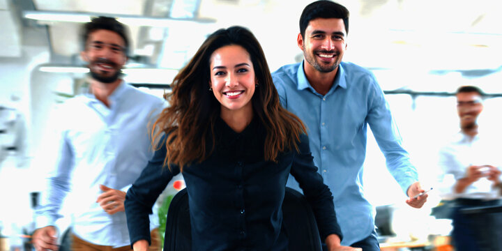 A diverse group of friendly young professionals laughing joyfully while playfully riding office chairs during a fun break in a bright modern workplace.