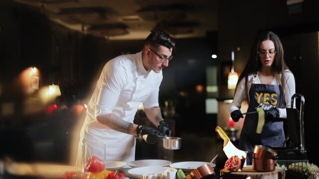 Male chef in white jacket and glasses leans over prep counter spooning sauce from small saucepan into white serving bowls while wearing black gloves, female assistant in dark apron and glasses