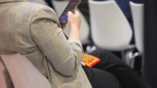 Person in checkered jacket sits, holds phone. Seat next to them has red patterned cloth. White chairs in background. Indoor setting with neutral lighting. Person focused on device