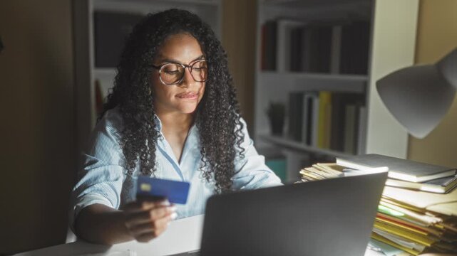 Woman holding creditcard and typing on laptop in a home office building at night; focused budgeting.