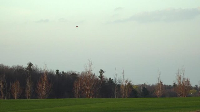 A powered paramotor flies high in a pale evening sky above bare deciduous trees and a vibrant green agricultural field.