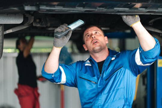 Ev car battery cells service Automotive mechanic inspecting vehicle underside with flashlight in repair garage, focused male technician wearing blue workwear and protective gloves