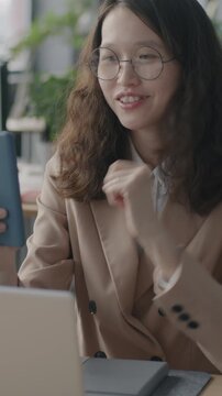 Young Asian woman making online video call with smartphone sitting at desk in office focused on professional communication. Business and virtual meeting concept.