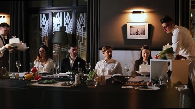 Two servers deliver covered cloche and bowls of fresh produce to four seated adults at long dining table in upscale interior, guests face camera with attentive expressions and formal attire