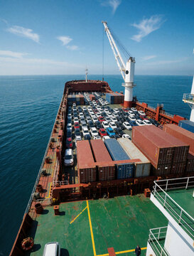 Large cargo ship carries hundreds of automobiles across the sea. Vessels deck packed with new cars, shipping containers stacked high. Global transport logistics, maritime industry.
