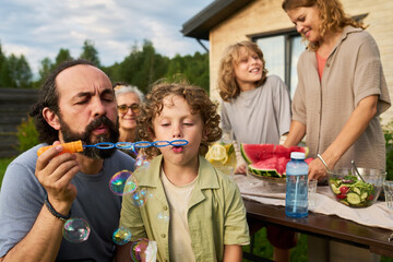 Fototapeta premium Caucasian father and male child blowing soap bubbles together during multigenerational outdoor family picnic. Ideal for family bonding advertising, parenting campaigns, summer leisure promotions
