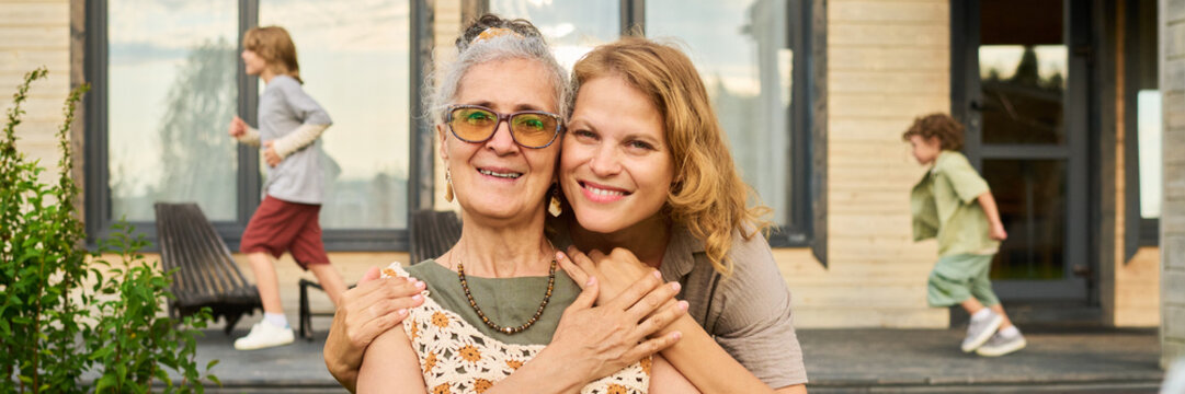 Portrait of smiling senior Caucasian woman and adult daughter embracing while children playing in backyard. Image suitable for multigenerational family insurance advertising