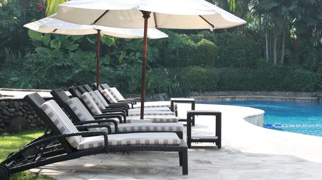 Rows of lounge chairs face a calm swimming pool. Large umbrellas shade the seating area. Lush greenery surrounds the poolside. Sunlight glints off the clear water