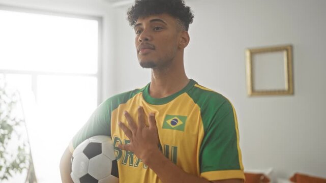 Man young brazilian wearing brazil jersey holding a soccer ball with hand placed on chest in building interior by a bright window; pride support.