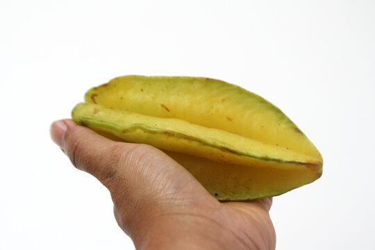 A hand holds a ripe yellow star fruit horizontally, showing its elongated shape, prominent ridges, and slightly green edges against a white background.