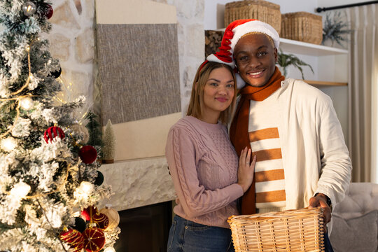 African American couple posing in living room by snowy tree holding wicker basket wearing Santa hat