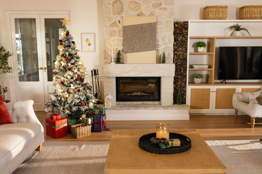 Flocked Christmas tree is glowing with warm lights in living room beside stone fireplace with gifts