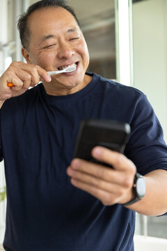 Senior Asian man in navy tee brushing with orange-toothbrush holding phone watch in bathroom mirror