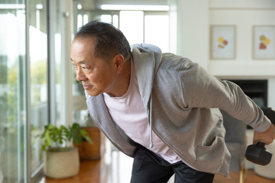 Senior Asian man leaning forward holding dumbbell in gray hoodie in living room by glass doors