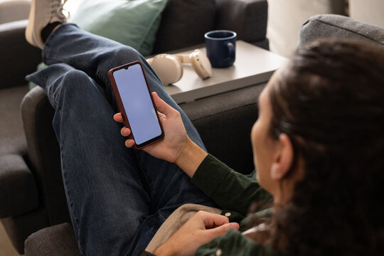 African American man reclining on gray sofa, holding phone with blank screen beside headphones tray