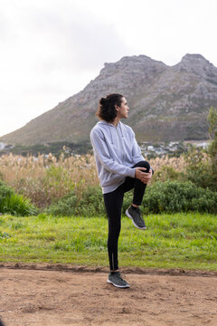 African American man stretching leg on trail at meadow, wearing gray hoodie, leggings