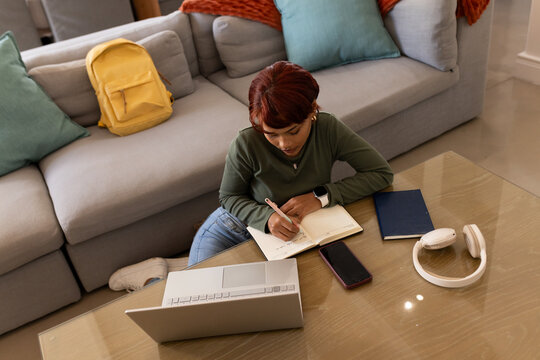 African woman sitting on floor at home writing in open notebook with laptop nearby
