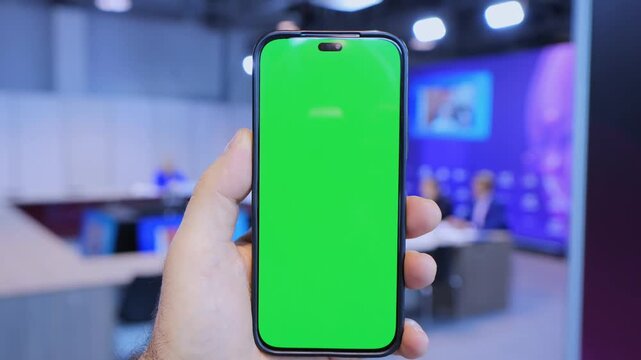 Hand holds smartphone with green screen. Background shows blurred tech event. Phone has black frame and no branding. Three keyframes show same scene. Indoor lighting illuminates hand and device