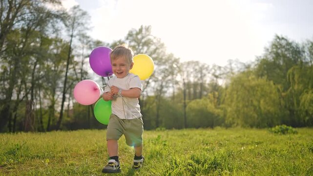 Boy stands smiling on summer grass holding balloon. Child looks happy in sunlight. Toddler grips balloon string tight. Outdoor air moves balloon. Bright colors fill field, balloon, child in frame