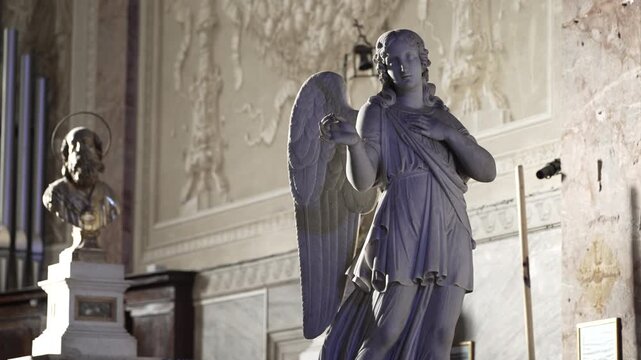 Marble angel statue stands in soft side light inside an old Italian Catholic church with carved walls and a bust in the background.