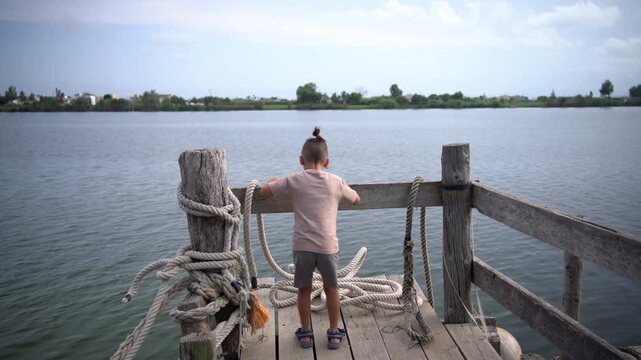 A boy walks onto a wooden dock with mooring ropes, leans on the railing, then climbs over it to look at the Ebro River in Tarragona, Spain