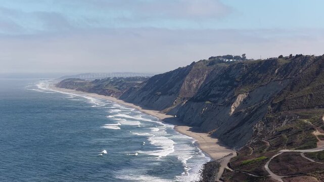 Aerial view of the coastline at Mussel Rock Park in Daly City, California, showing crashing waves on the sandy beach against dramatic cliffs under a hazy sky