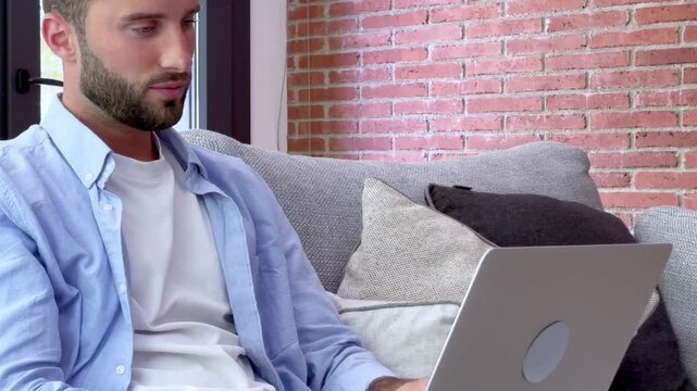 Thoughtful young man working on a laptop computer while sitting on a comfortable sofa at home, successfully solving a problem and feeling relieved while smiling and working remotely