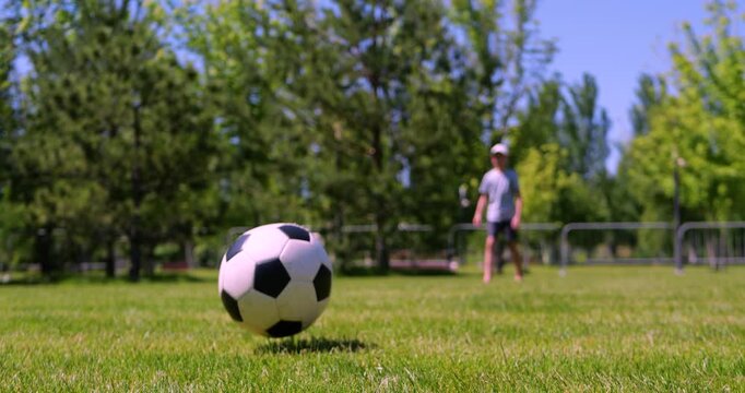 Barefoot child kicks soccer ball in sunny park as camera moves backward and focus shifts from child to rolling ball close up. Cinematic outdoor childhood and casual summer play.