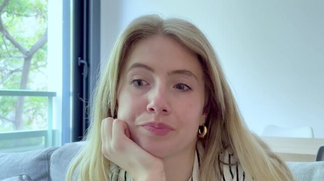 Frustrated blonde woman sitting on the couch in her living room, displaying boredom and impatience while waiting for something, with apathetic and tired expressions on her face