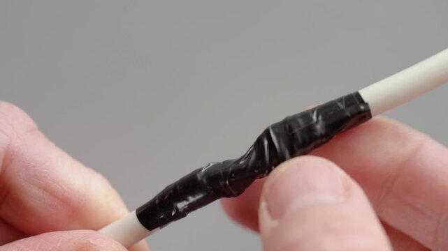 Closeup hands inspecting taped wire, manual technician checking how black vinyl electrical tape adheres to white insulated cable, fingertips pressing and running along wrap, studio